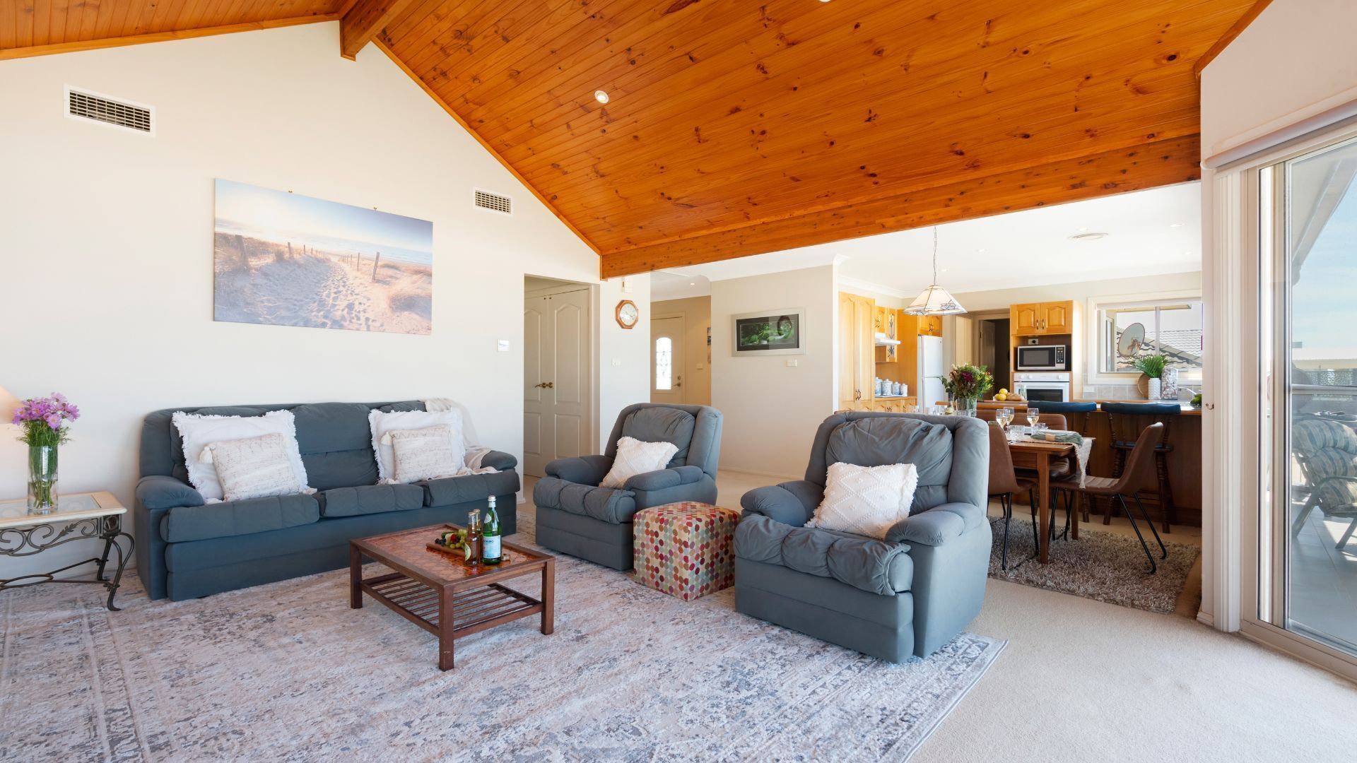 A spacious living room with a high wooden ceiling, a cozy sofa set, a coffee table, and a view of the kitchen and balcony.