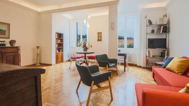 A spacious living room with a wooden floor, a red sofa, two black chairs, a dining table with chairs, and a bookshelf.