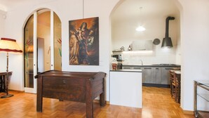 A room with a wooden floor, a dark wooden table, a painting, a kitchen area with cabinets and a stove, and a lamp.