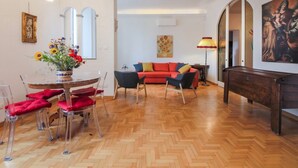A spacious living room with a wooden herringbone floor, a round glass dining table with red cushions, a red sofa with colorful cushions, and a dark wooden bench.