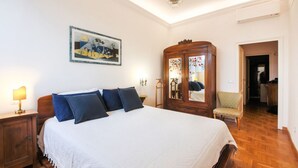 A bedroom with a bed, a wooden wardrobe, a nightstand, a lamp, a chair, and a painting on the wall.