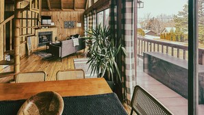 Un intérieur de chalet chaleureux avec un escalier en bois, une cheminée, un coin repas et un balcon offrant une vue sur le paysage environnant.