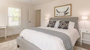 Quarto com cama, mesas de cabeceira, ventilador no teto, janela com persianas e um quadro na parede.