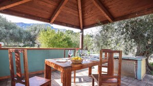 A wooden table set for two with a bowl of fruit, wine glasses, and a bottle on a patio with a view of mountains and trees. - 2 bedroom awesome home in Altavilla Milicia (Altavilla Milicia)