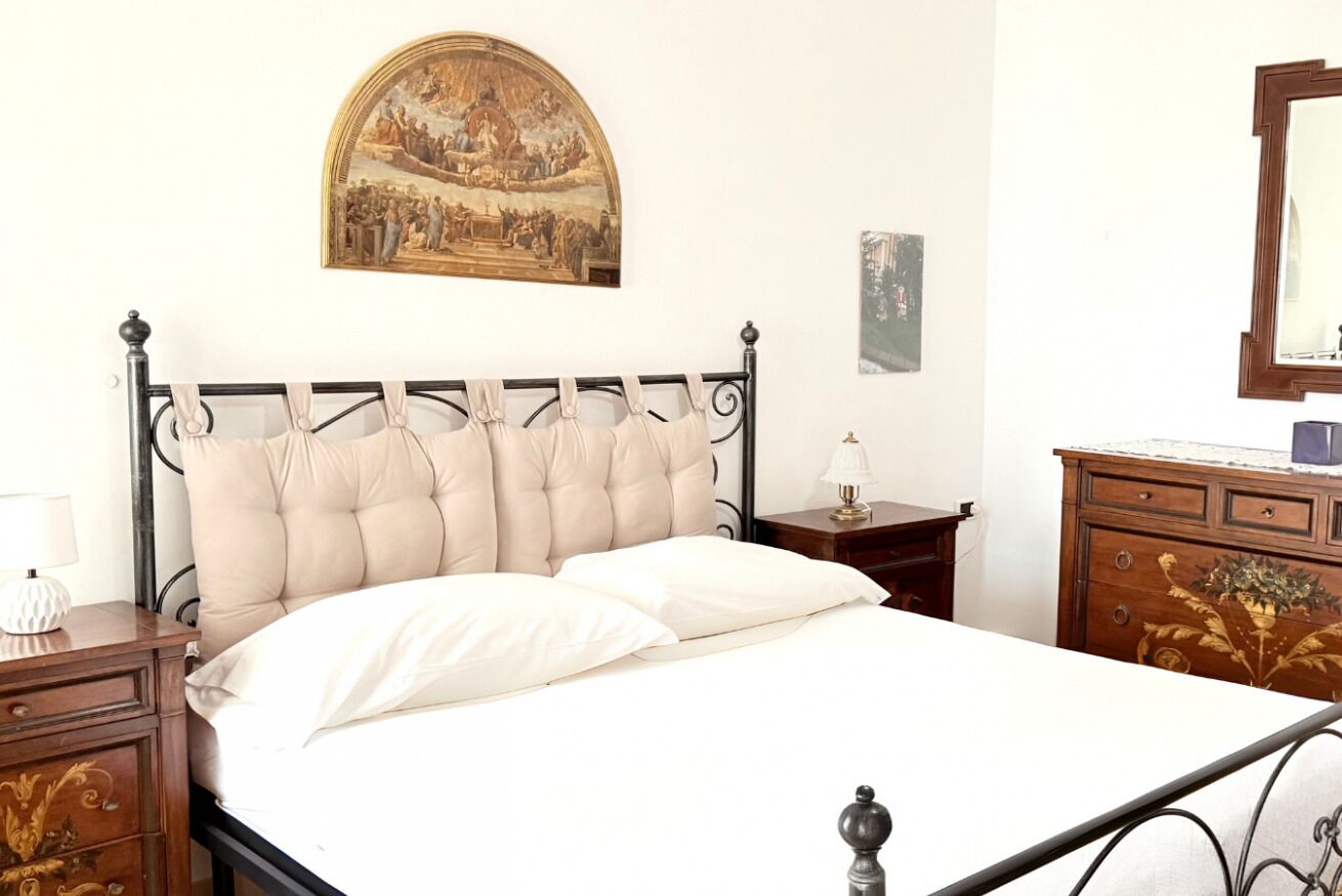 Un letto con una testiera, un comodino con una lampada, una cassettiera e uno specchio appesi al muro, oltre a un quadro incorniciato.