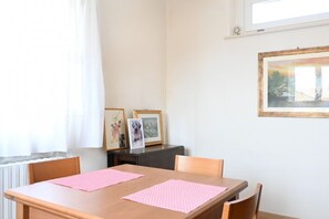 Un coin repas avec une table en bois, deux sets de table roses et des tableaux encadrés posés sur une petite table d’appoint de couleur foncée.