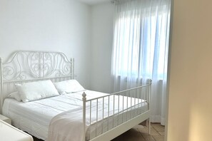 A white metal bed with a headboard, white bedding, and a window with sheer curtains.
