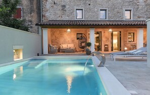 Un espace piscine avec un bâtiment en pierre en arrière-plan, des sièges d’extérieur et un fauteuil suspendu.
