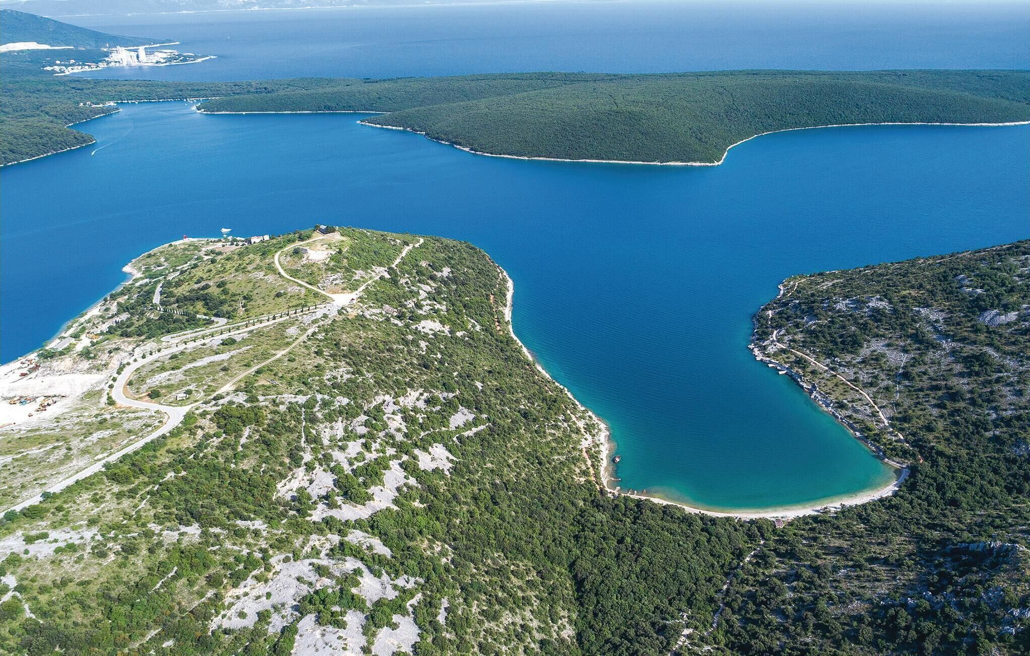An aerial view of a coastal area with a winding road, dense greenery, and a clear blue sea.