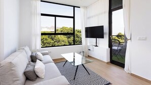 Una sala moderna con un sofá, una mesa de centro de vidrio, un televisor de pantalla plana y un ventanal con vistas a zonas verdes.