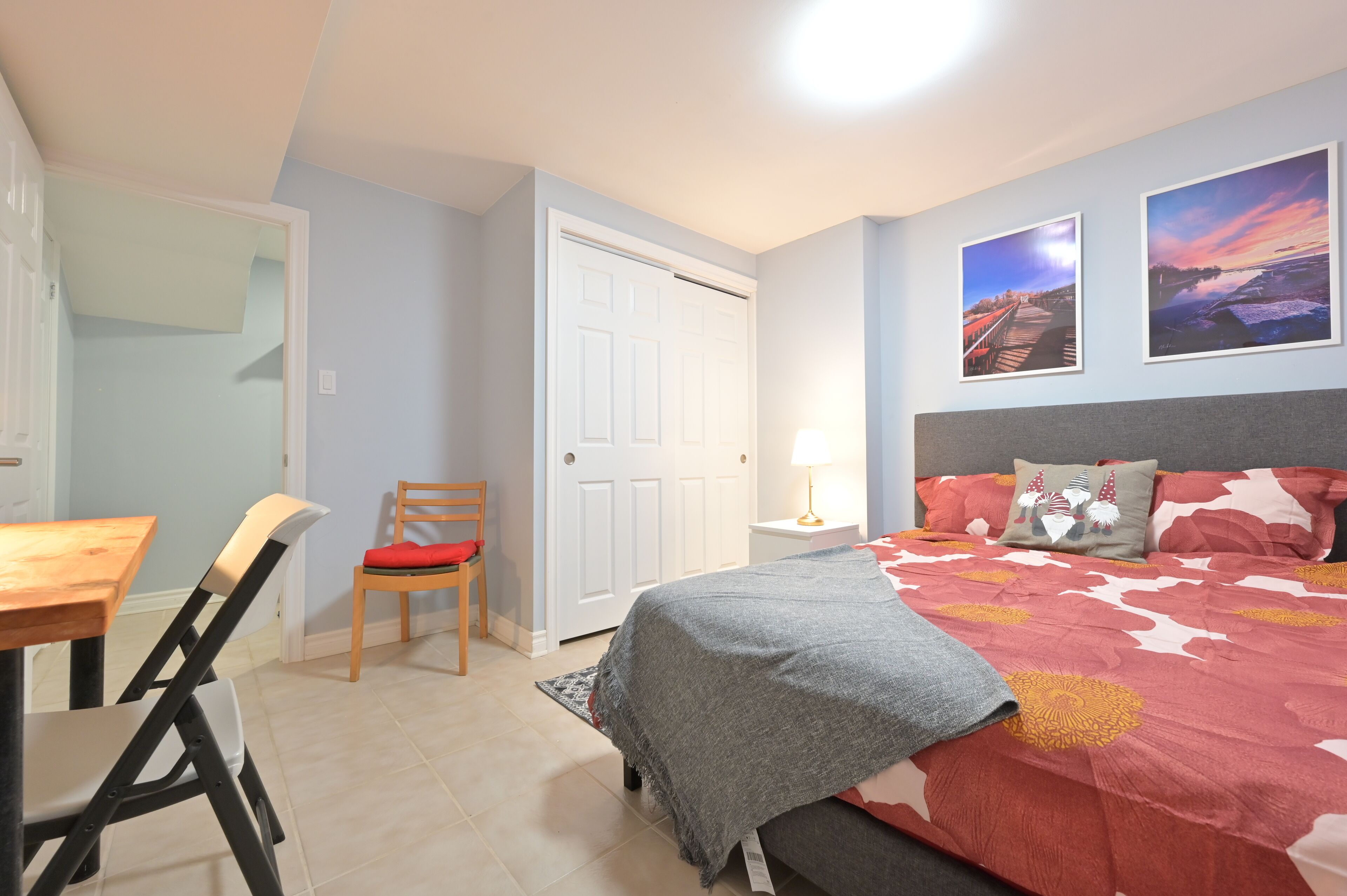 A bedroom with a bed, a desk, and a chair. There are two paintings on the wall.