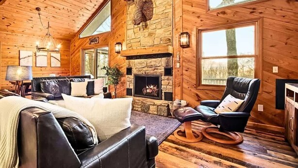 A log cabin interior with a stone fireplace, leather furniture, and a large window.
