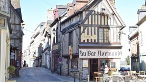 Eine schmale Gasse mit traditionellen Fachwerkhäusern, einem Café namens 'Au Bar Normand' und Kopfsteinpflaster.