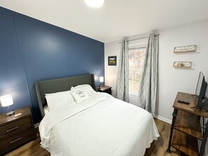 Quarto com cama, mesa de cabeceira, escrivaninha e janela com cortinas.