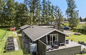 En træhytte med en overdækket terrasse, en lille have og en bil parkeret i nærheden.
