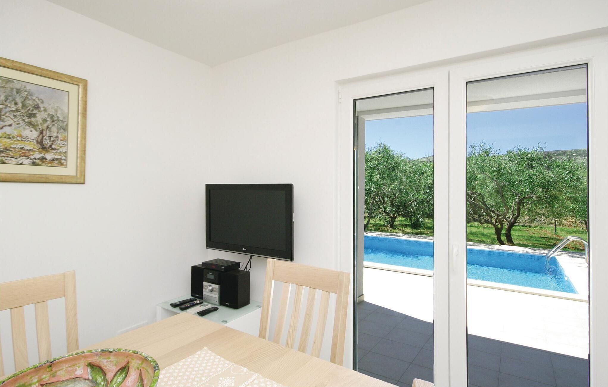 A room with a TV, a table, chairs, and a view of a swimming pool.