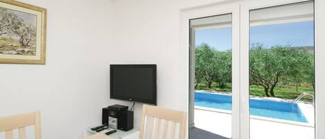 A room with a TV, a table, chairs, and a view of a swimming pool.