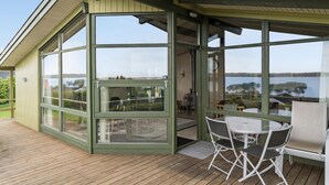 Ein überdachter Außenbereich mit Glasfassade, einem Tisch mit Stühlen und Blick auf einen See.