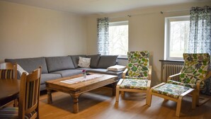 Ein Wohnzimmer mit einer Couch, zwei Sesseln, einem Holztisch und einem Esstisch mit Stühlen.