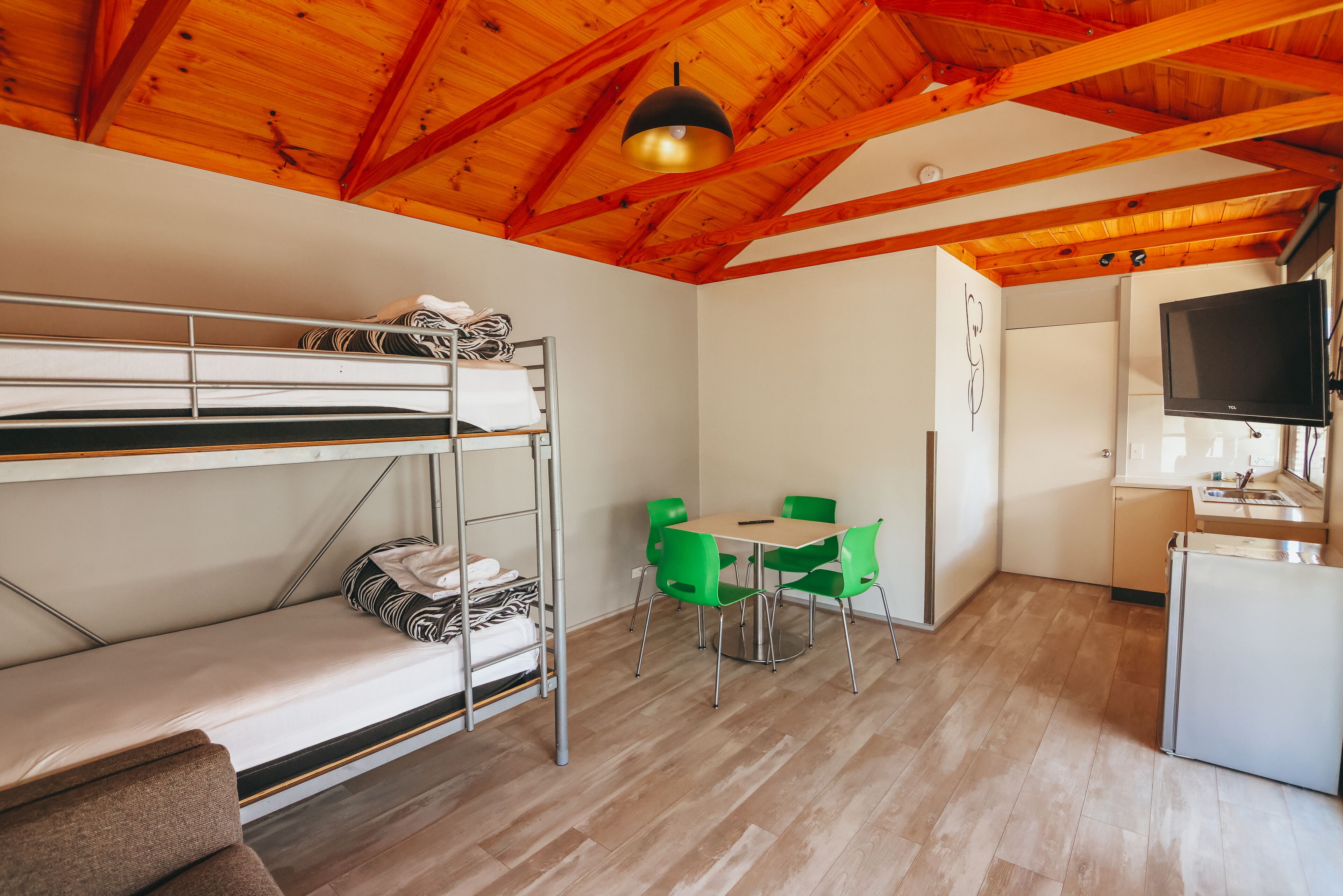 Un lit superposé, une table à manger avec des chaises, un coin cuisine avec un téléviseur et des poutres en bois au plafond.