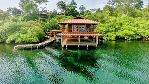 Une maison en bois avec un toit rouge, construite sur une plateforme flottante au milieu d’une étendue d’eau, et entourée d’une végétation luxuriante.