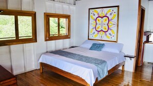 Une chambre avec un lit en bois, des murs blancs, une grande fenêtre et un tableau abstrait aux couleurs vives.