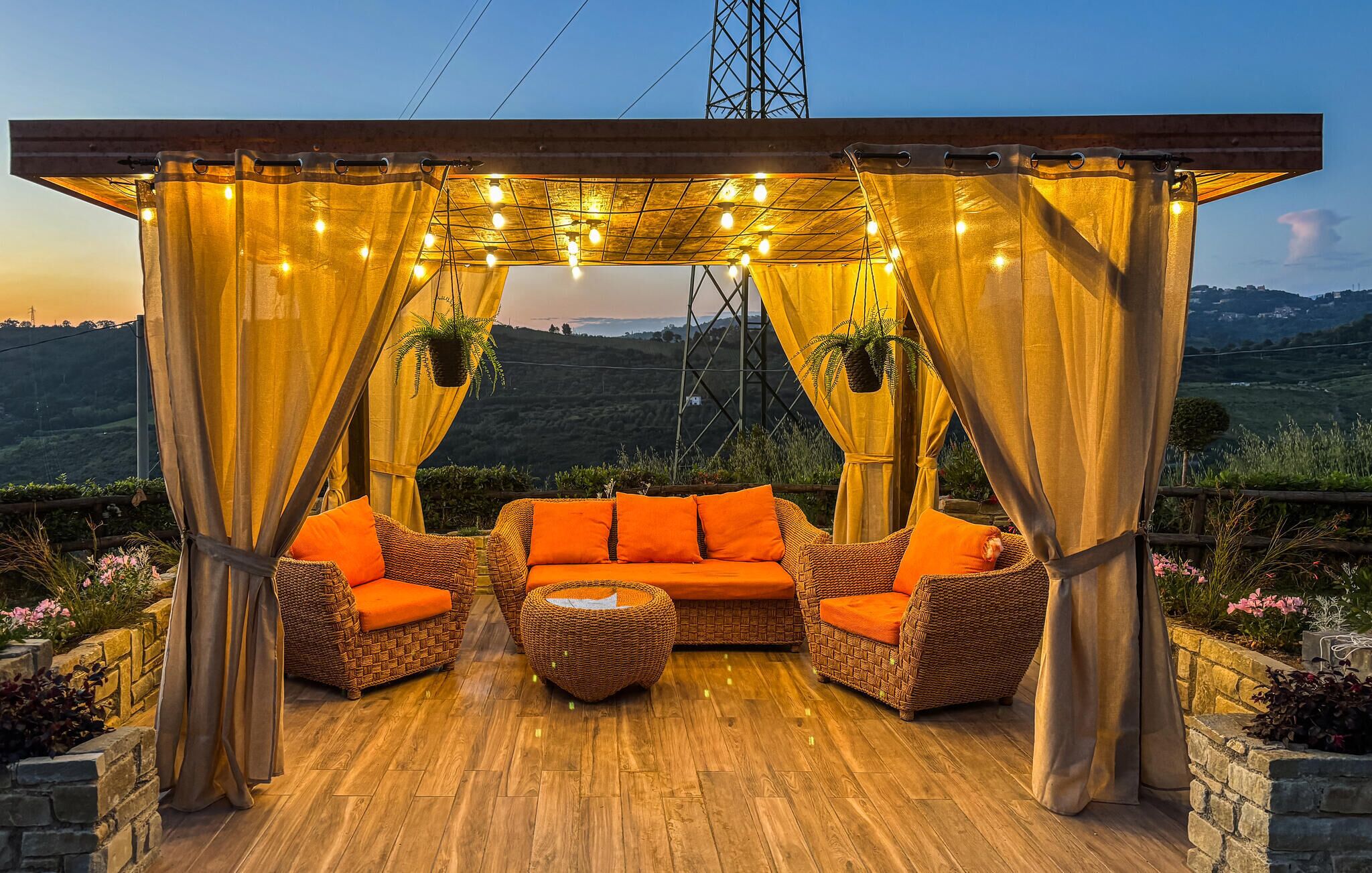 Un espace extérieur aménagé avec des meubles en osier et des coussins orange, sous une tonnelle illuminée de guirlandes lumineuses, offrant une vue sur un paysage vallonné.