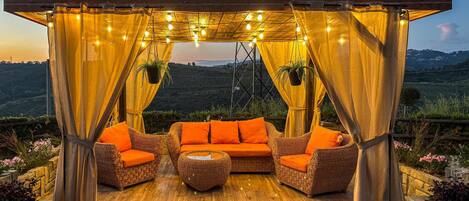 Un espace extérieur aménagé avec des meubles en osier et des coussins orange, sous une tonnelle illuminée de guirlandes lumineuses, offrant une vue sur un paysage vallonné.
