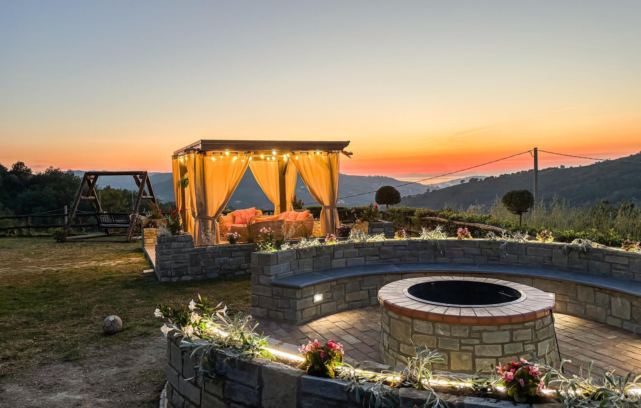 A gazebo with string lights, a stone fire pit, and a view of the sunset over a hilly landscape.