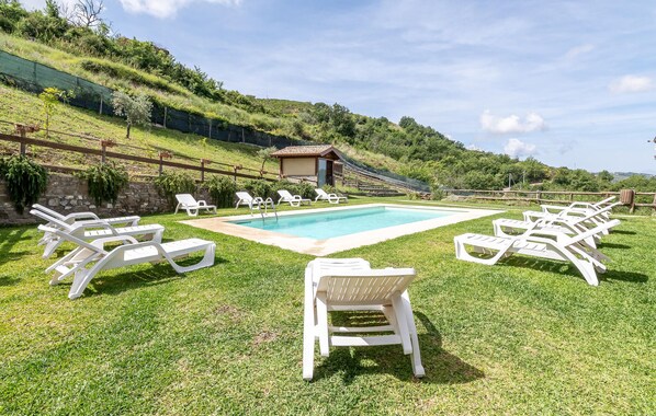 Zona de piscina amb gandules blanques disposades al seu voltant, un edifici petit al fons i una vessant de pendent coberta d'herba.