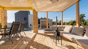 A covered outdoor area with stone flooring, a pool, a dining setup, and a lounge with a sofa and chairs.