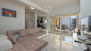 A modern living room with a sectional sofa, a dining area with outdoor seating, and a kitchen visible through an open door.