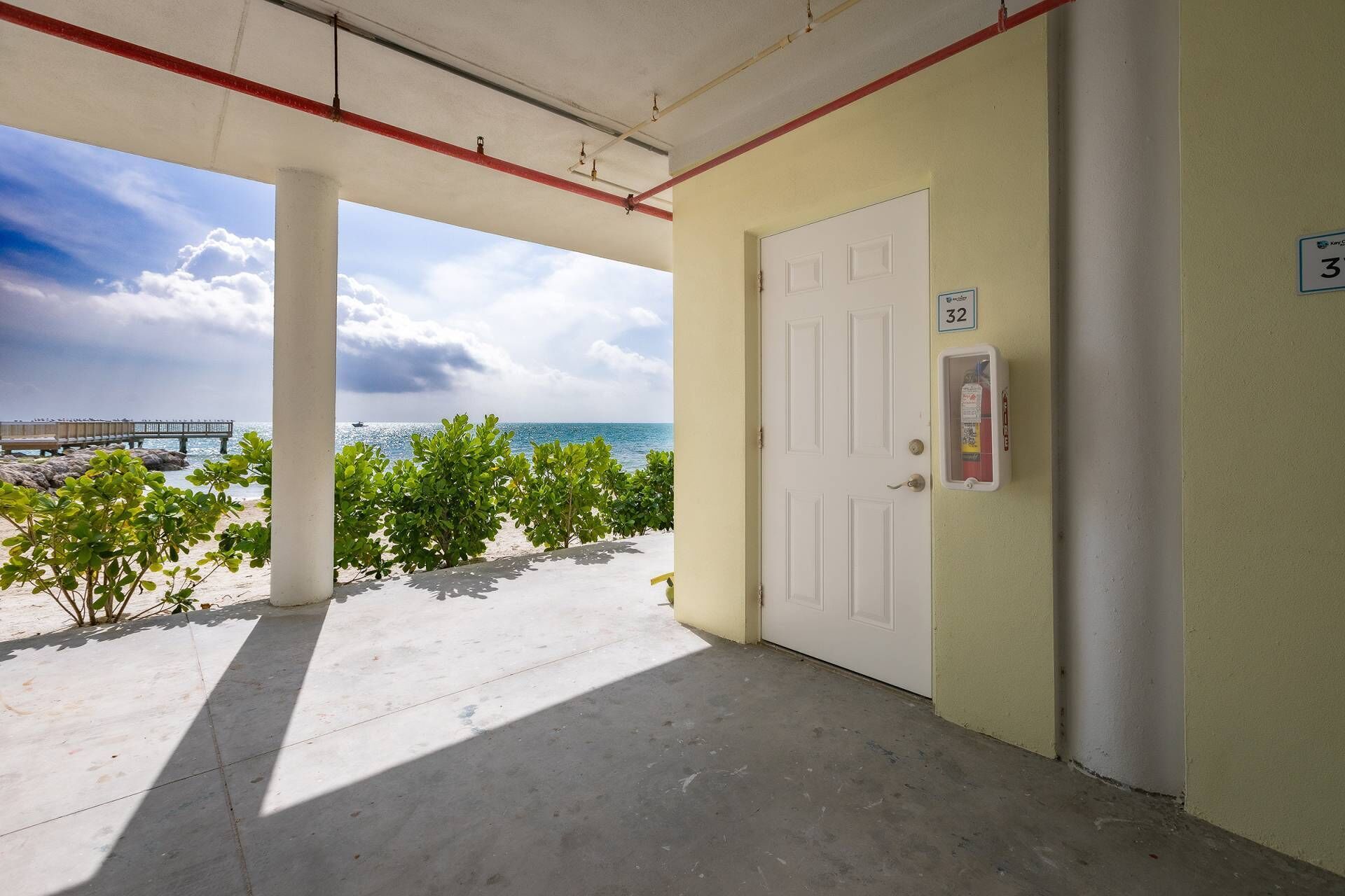 Un espace extérieur abrité doté d’un sol en béton, d’une porte blanche et d’un extincteur, offrant une vue sur la plage et l’océan.