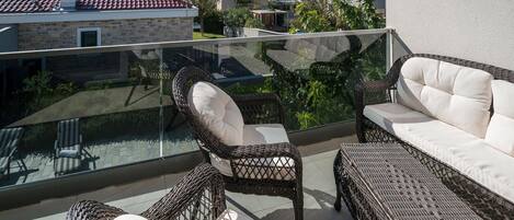 A balcony with wicker furniture, a glass balustrade, and a view of the sea and neighboring buildings.