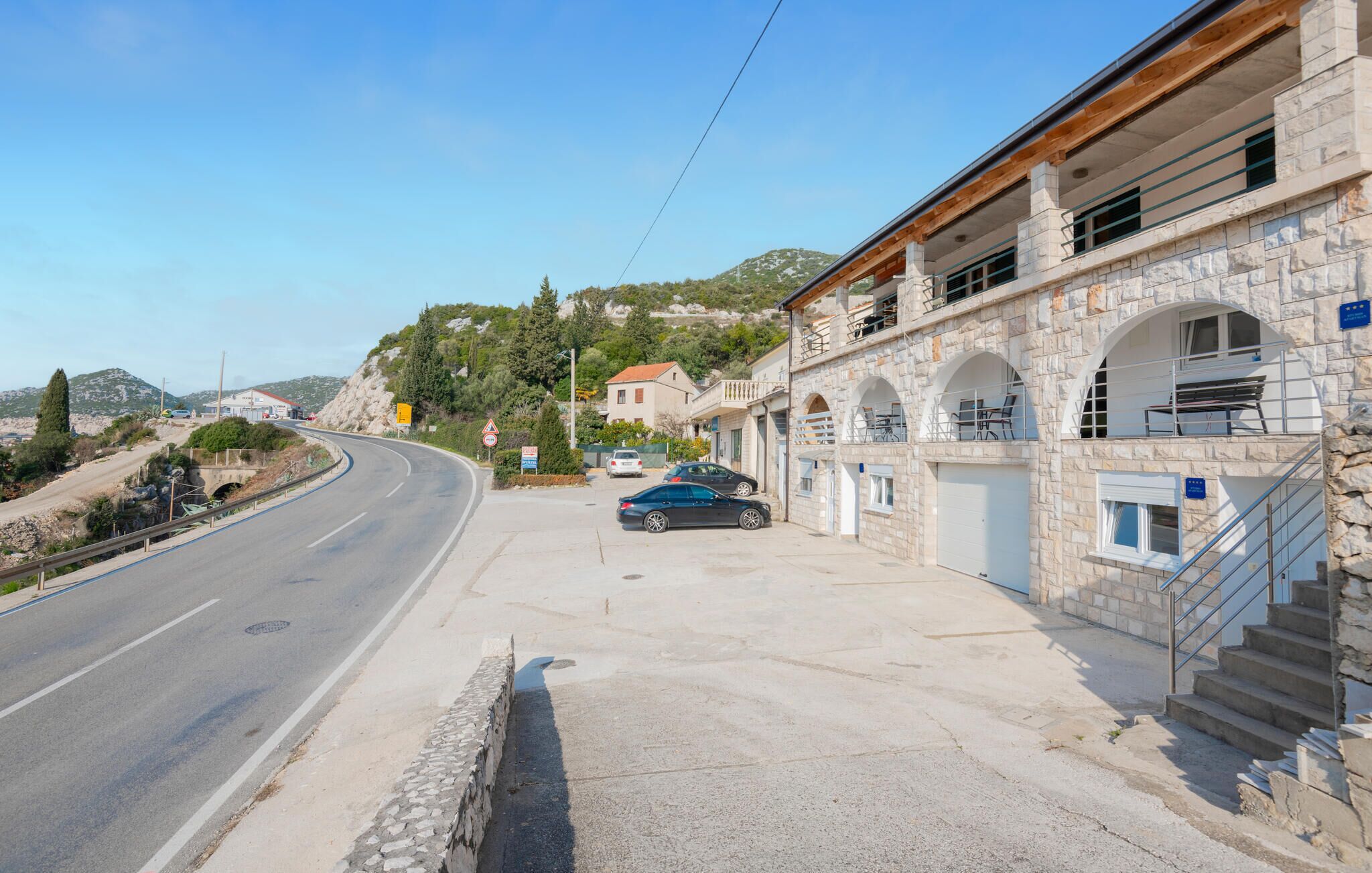 A stone building with a staircase leading to an upper level, a parked car, and a curved road with a guardrail.
