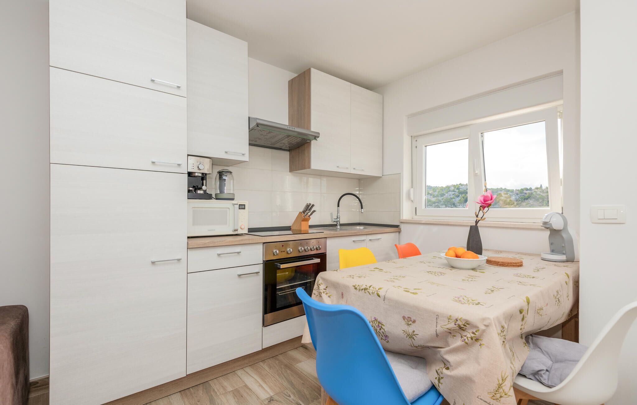 Een kleine keuken met witte kasten, een ingebouwde oven, een magnetron en een koffiezetapparaat. Er staat een eettafel met een tafelkleed met bloemenmotief en een vaas met bloemen.