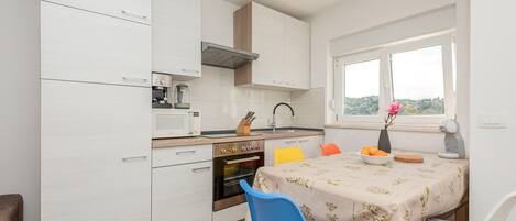 Een kleine keuken met witte kasten, een ingebouwde oven, een magnetron en een koffiezetapparaat. Er staat een eettafel met een tafelkleed met bloemenmotief en een vaas met bloemen.
