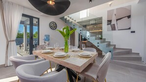 Un coin repas avec une table en bois dressée pour six personnes, entourée de chaises blanches aux pieds dorés. On aperçoit un grand distributeur d’eau à l’arrière-plan.