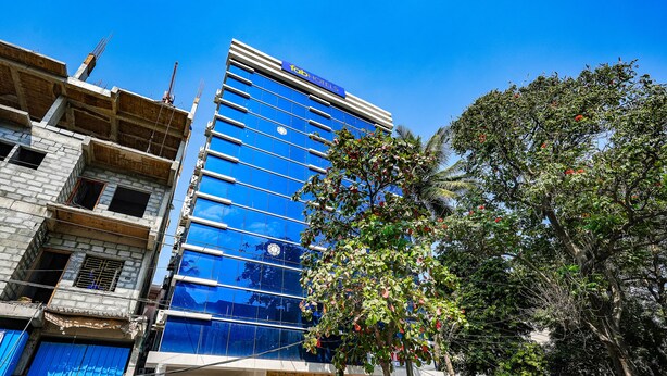 A modern hotel with a glass facade, a sign for 'HOTEL 24 PLUS', and a tree in the foreground. - Fabexpress 24 Plus (Bengaluru)