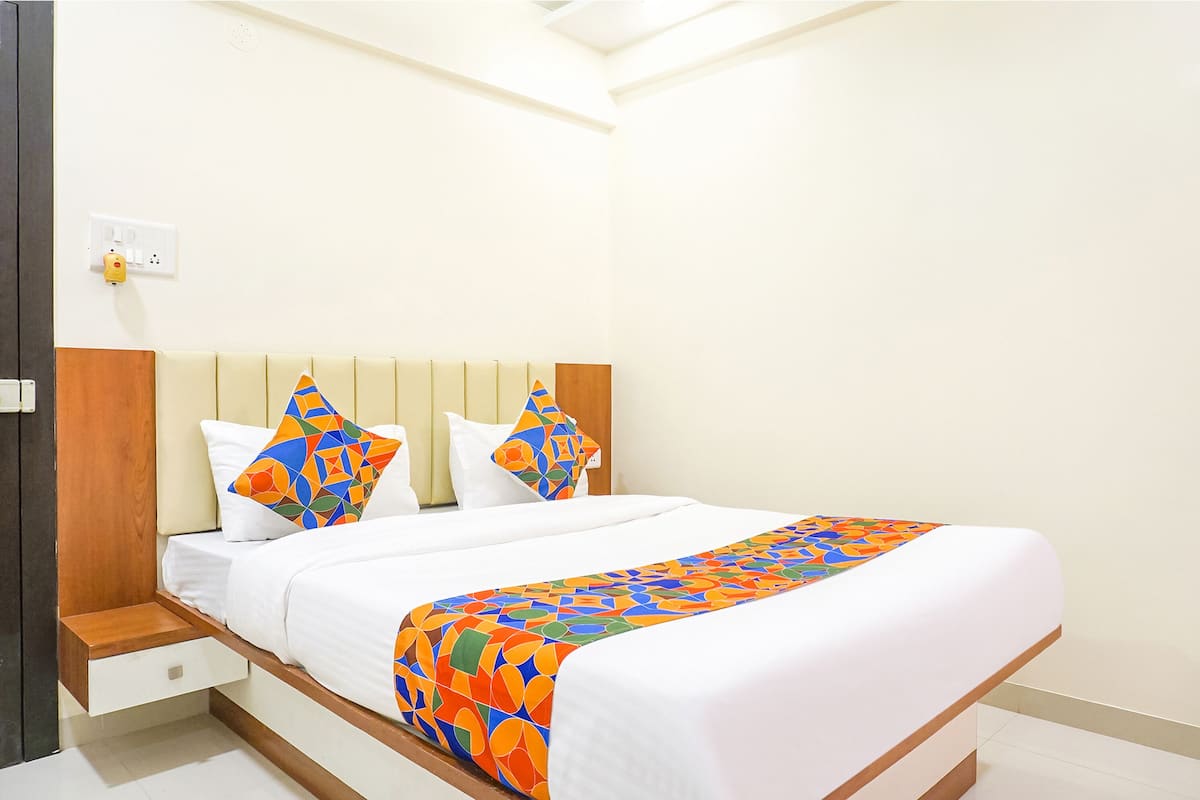 Uma cama bem arrumada com um edredom colorido e duas almofadas, uma mesinha de cabeceira de madeira e uma parede branca com uma tomada.