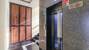 A hallway with a wooden door, a stairway, and a glass elevator door. There are signs indicating a fire exit and a no-smoking zone. - Fabexpress Grand Inn (Lucknow)