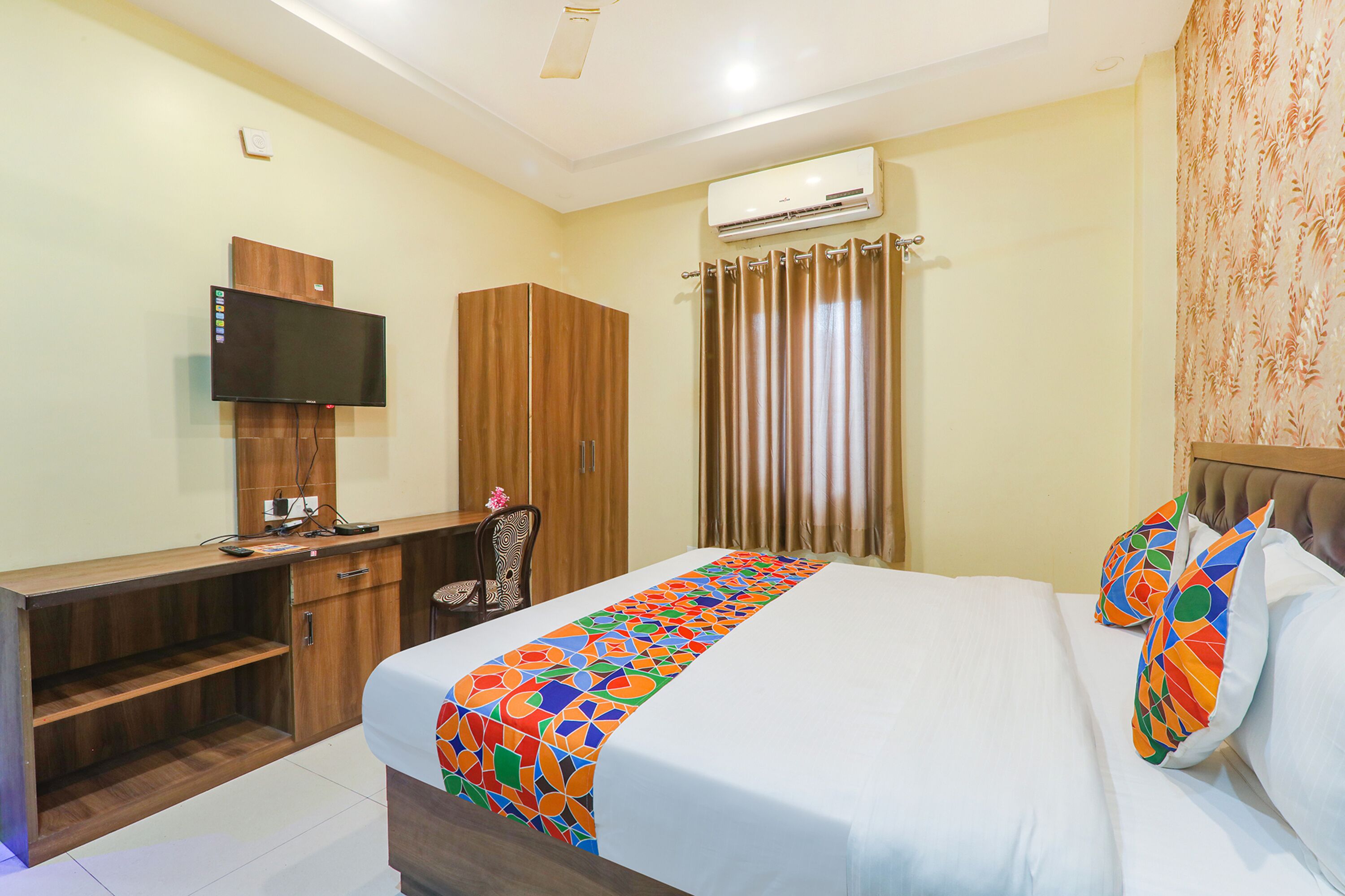 A hotel room with a bed, a desk with a TV, a wardrobe, and an air conditioning unit.
