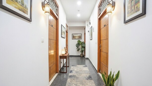 A hallway with wooden doors, wall-mounted lights, framed pictures, a potted plant, and a small table with a lamp. - Fabhotel Prime Residency (New Delhi)