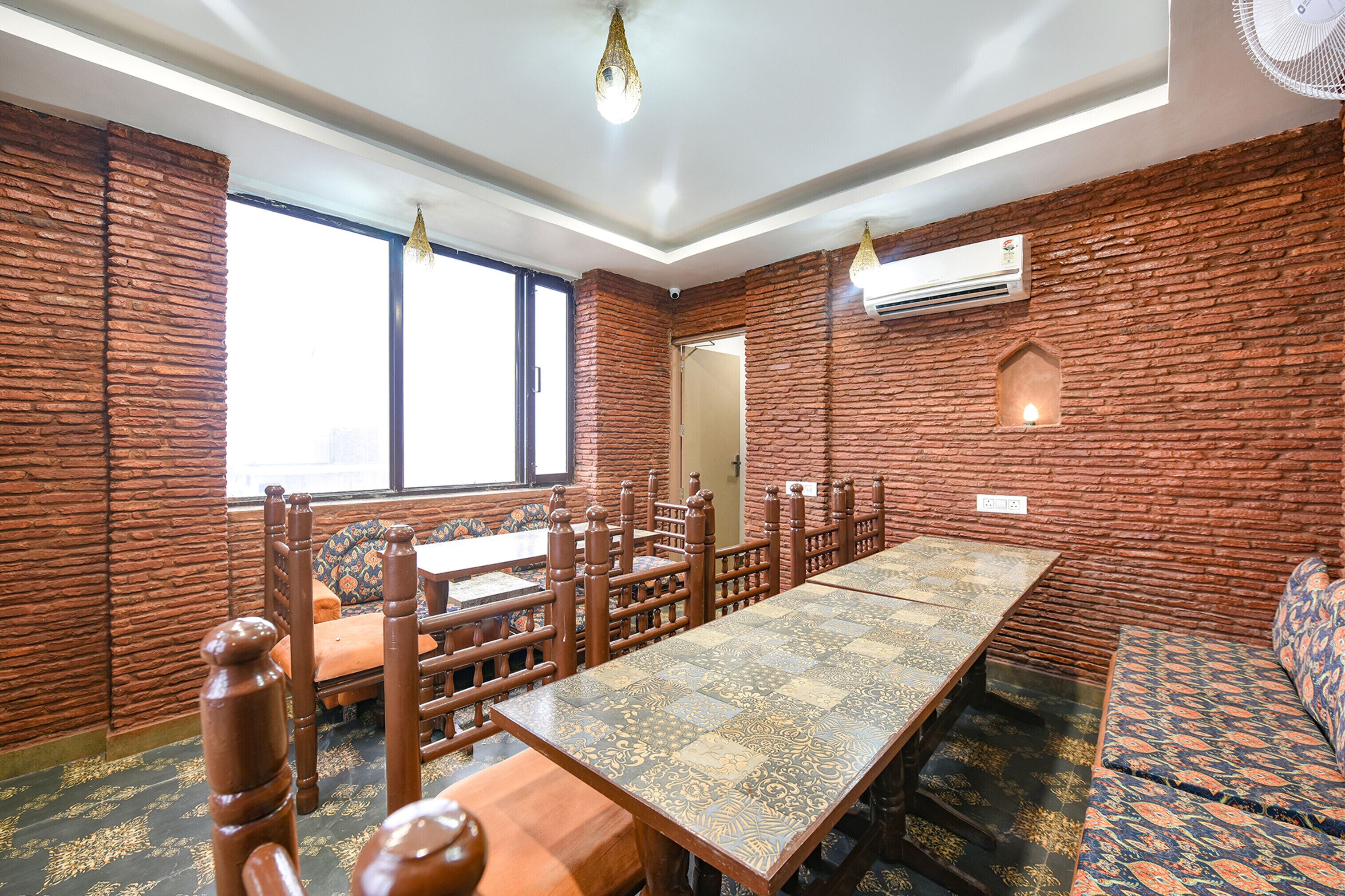 Uma sala de jantar com uma mesa comprida, cadeiras e bancos de madeira. Há janelas, um aparelho de ar condicionado e um lustre.