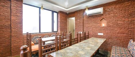 Uma sala de jantar com uma mesa comprida, cadeiras e bancos de madeira. Há janelas, um aparelho de ar condicionado e um lustre.