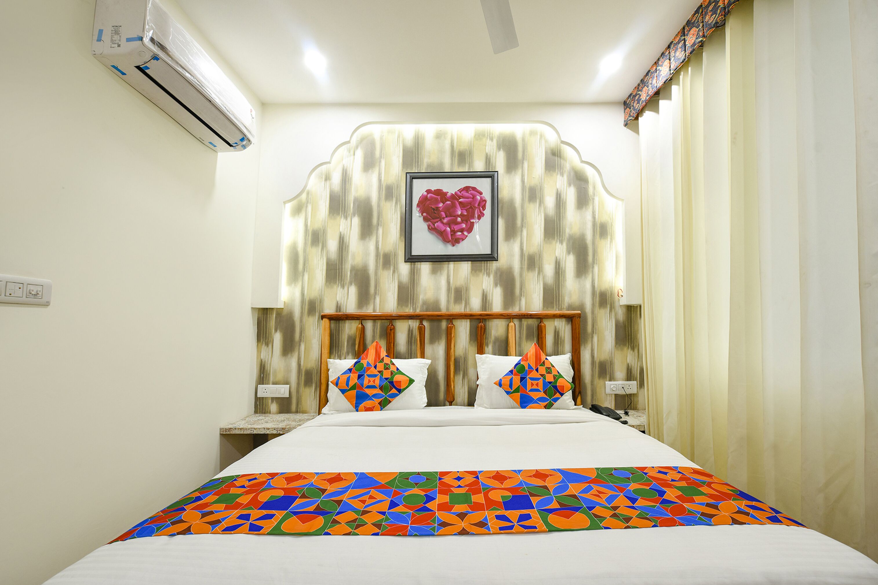 A hotel room with a bed, a headboard with a heart-shaped decoration, and a patterned bedspread.