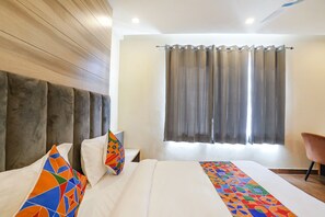 A hotel room with a wooden headboard, a large bed with colorful pillows, a window with curtains, and a chair. - Fabhotel Js Pearl (Dehradun)