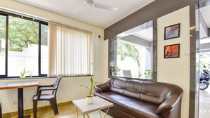 A modern living room with a brown leather sofa, a wooden table with a potted plant, a chair, and large windows offering a view of greenery outside.