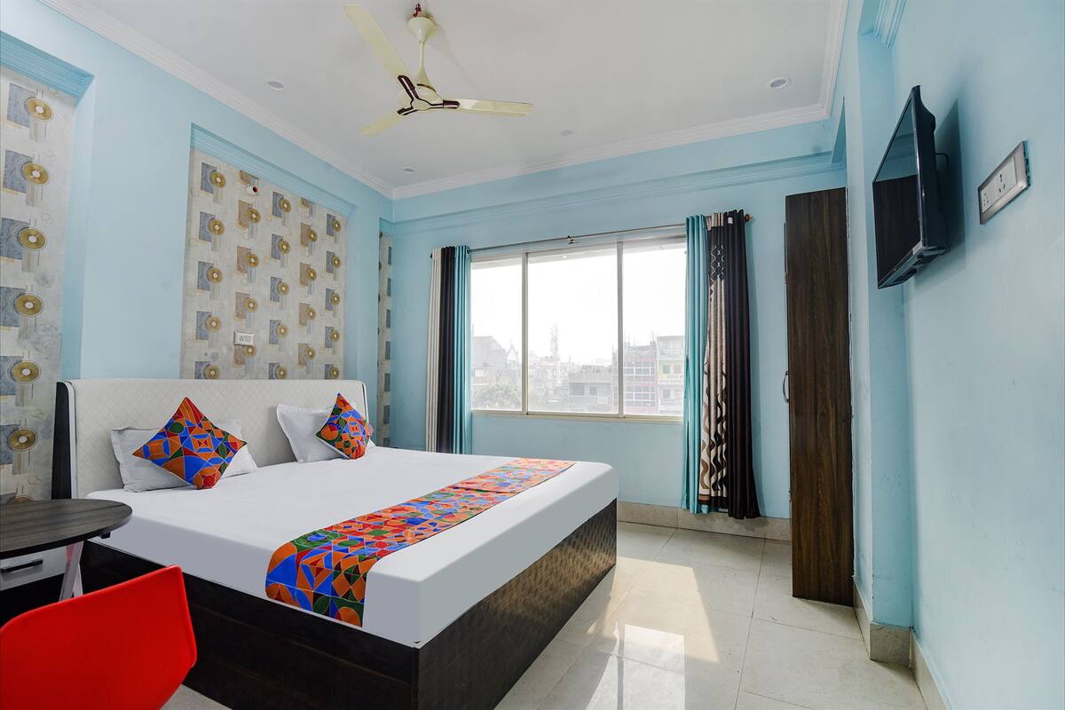 A hotel room with a bed, a TV, a ceiling fan, a window with curtains, and a red chair.