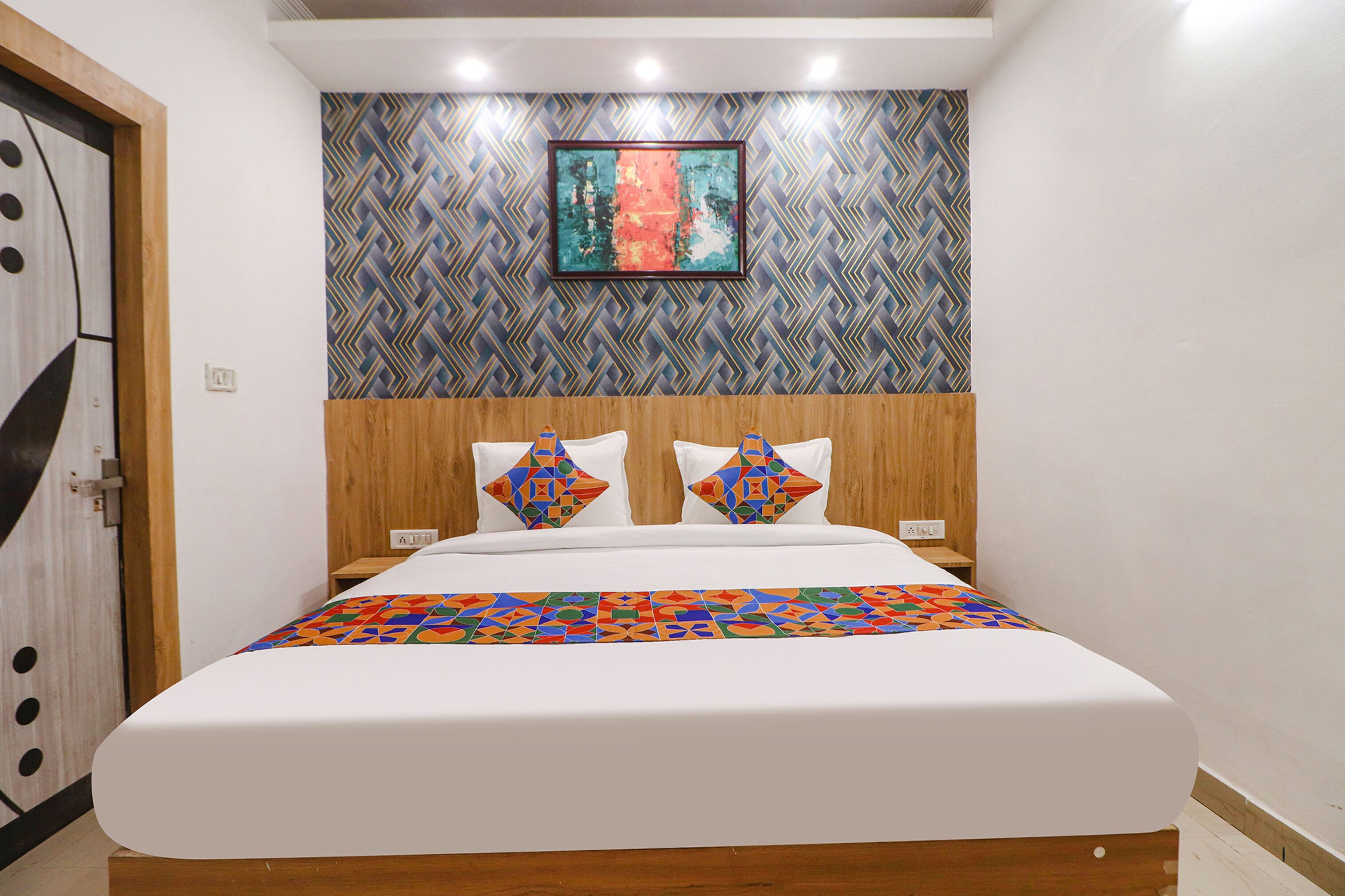 A hotel room with a large bed, a wooden headboard, a colorful geometric-patterned bedspread, and a wall with a geometric-patterned wallpaper.